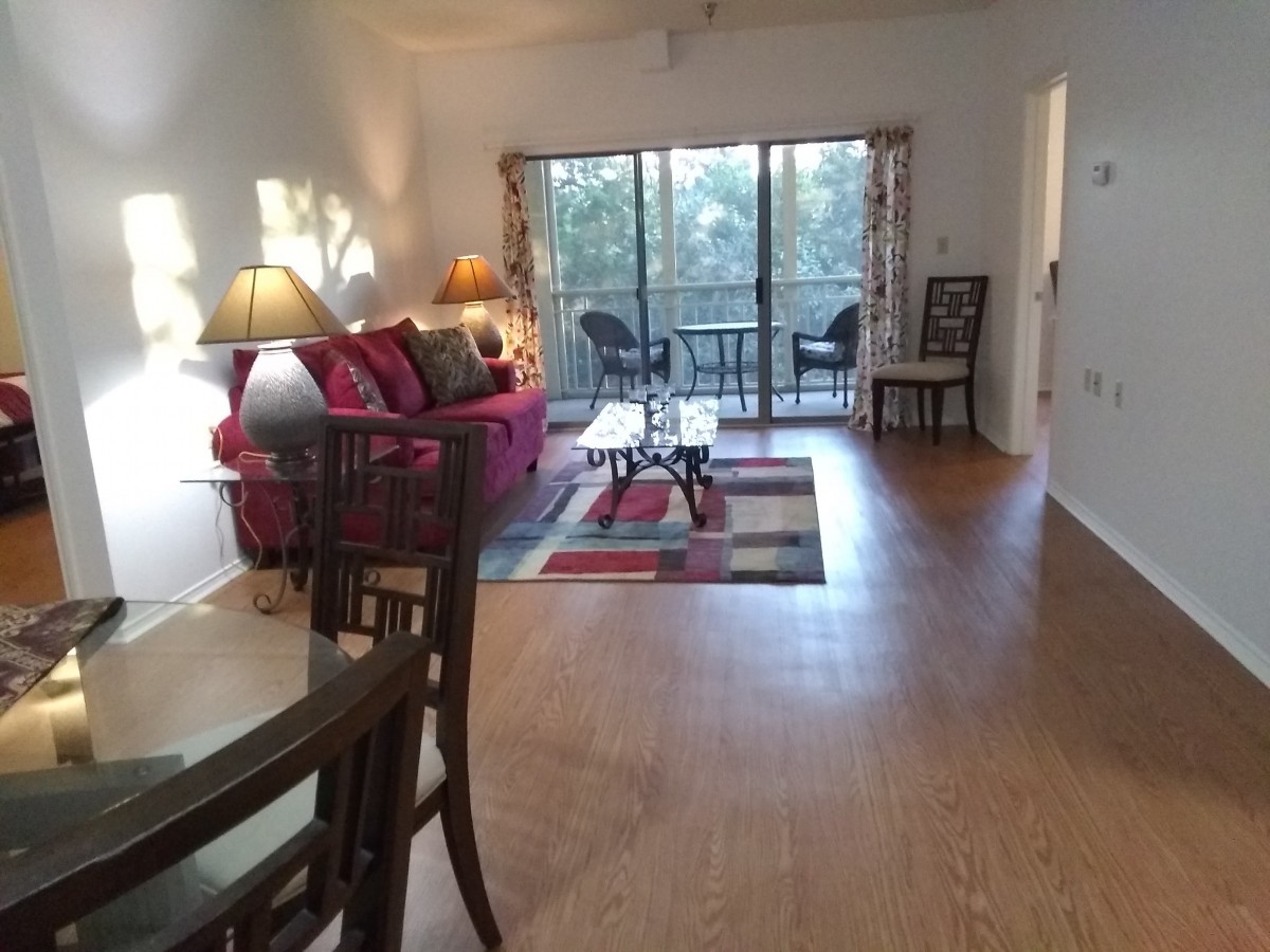 A cozy living room with a red sofa, coffee table, and dining area, featuring wood flooring and sliding glass doors leading to a balcony.