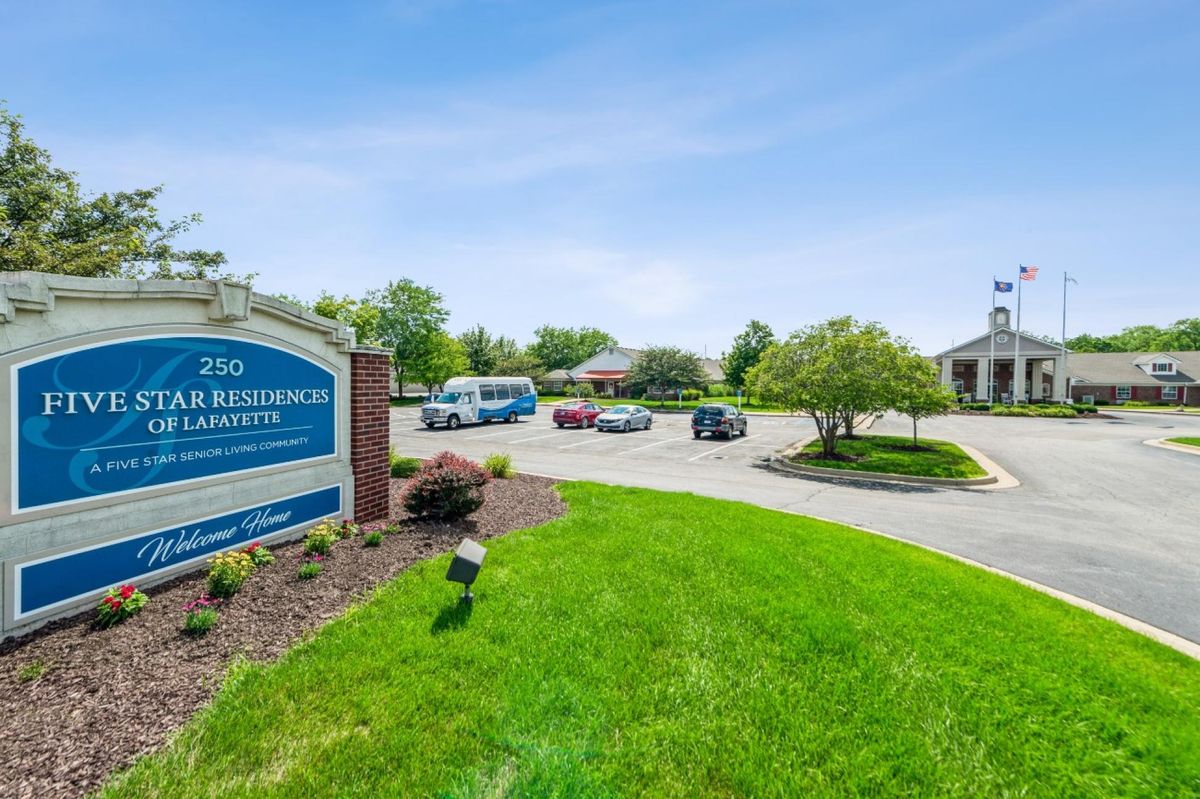 A view of an office complex with a sign, parking lot, and surrounding greenery on a clear day.