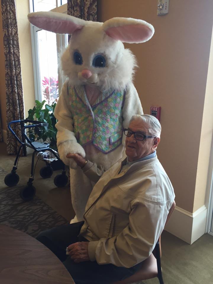 An elderly man sitting next to a person in an Easter Bunny costume indoors.