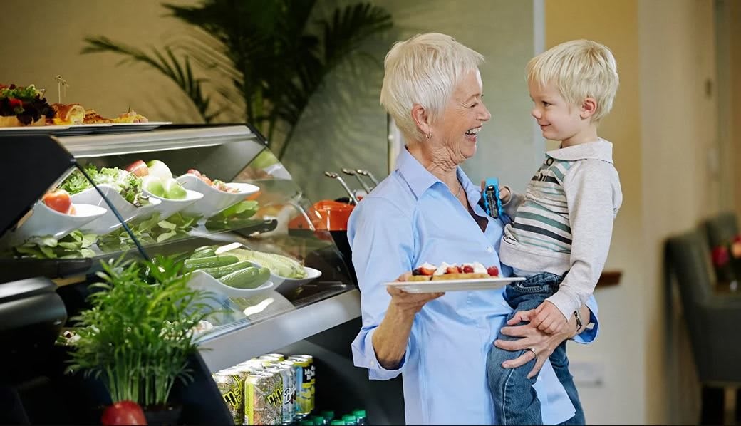 A grandmother happily holds her grandson while carrying a plate of food indoors.