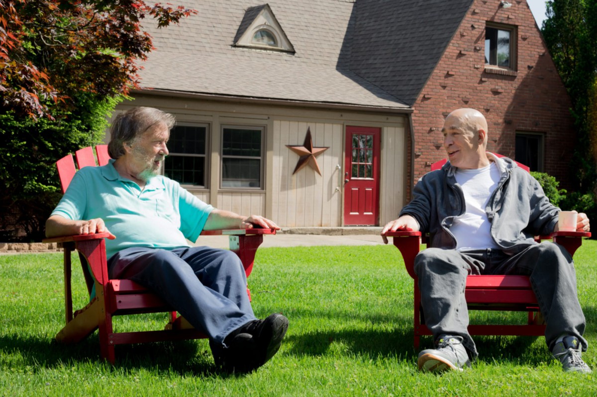 Two individuals sitting on red chairs, conversing on a grassy lawn in front of a house.