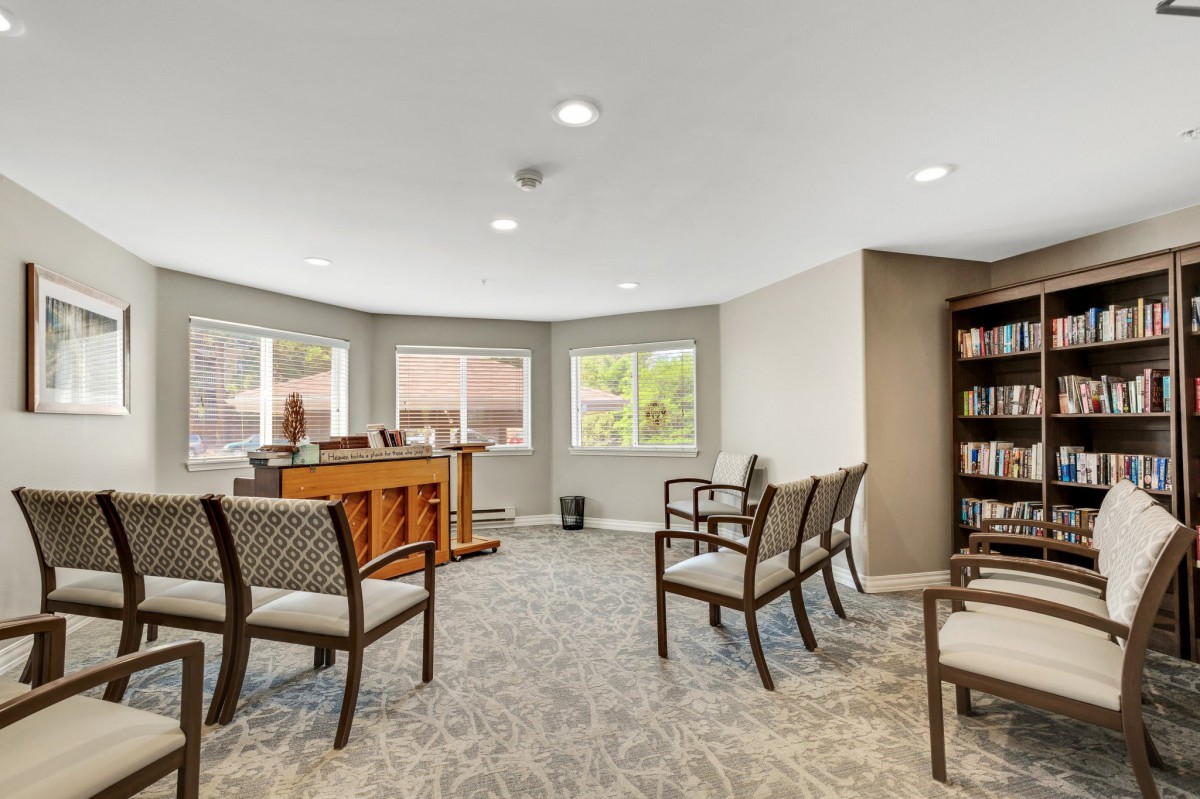 A well-lit room with several chairs and a wooden desk, featuring large windows and a bookshelf.