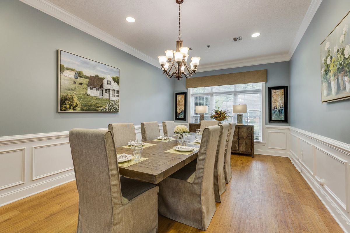 An elegantly decorated dining room with a long table and upholstered chairs, featuring a chandelier and artwork on the walls.