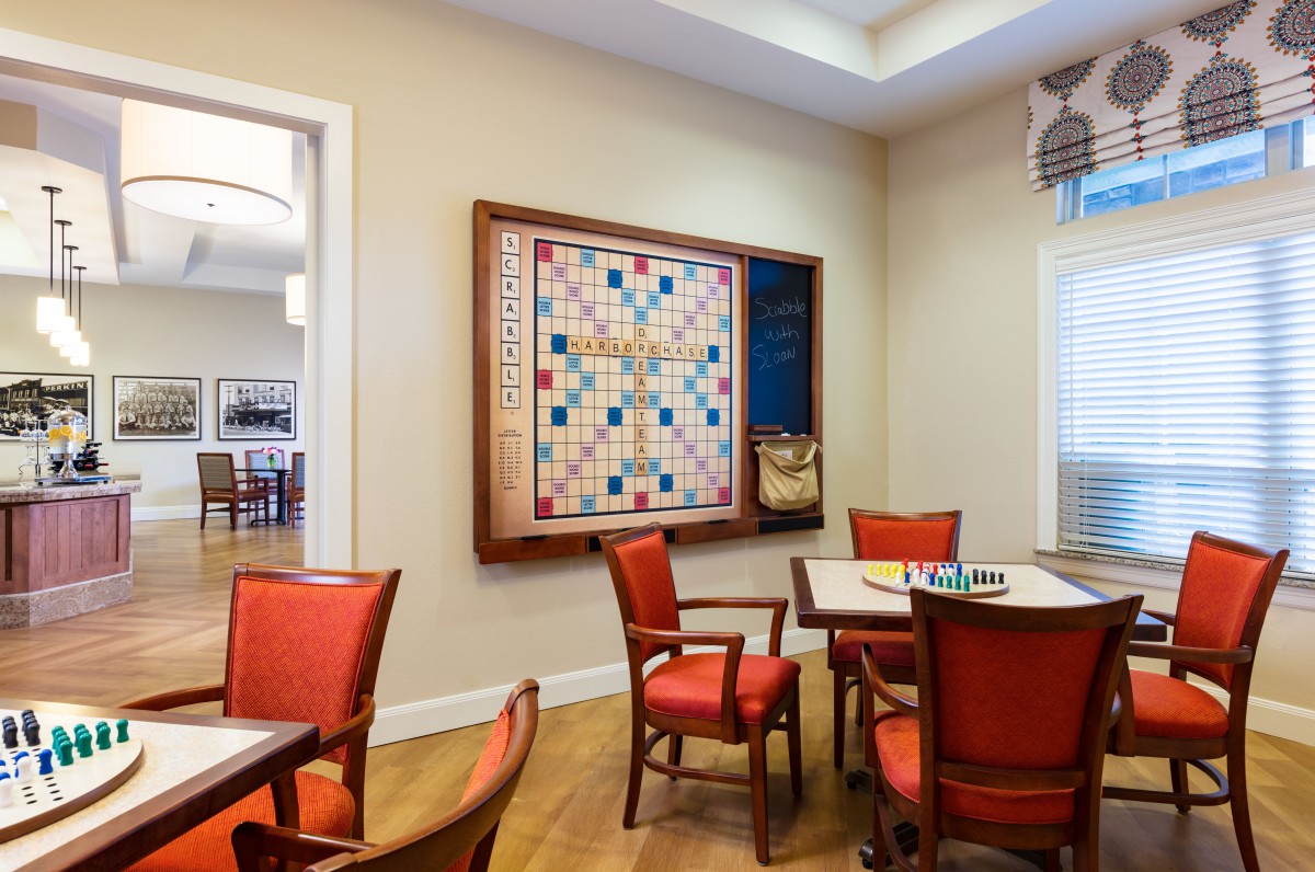 A cozy game room featuring a large Scrabble board on the wall, surrounded by red chairs and tables for gameplay.