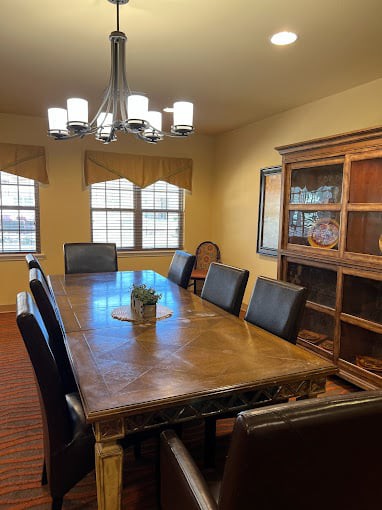 A spacious dining room with a large wooden table, surrounded by chairs and illuminated by a chandelier, featuring a cabinet and multiple windows.