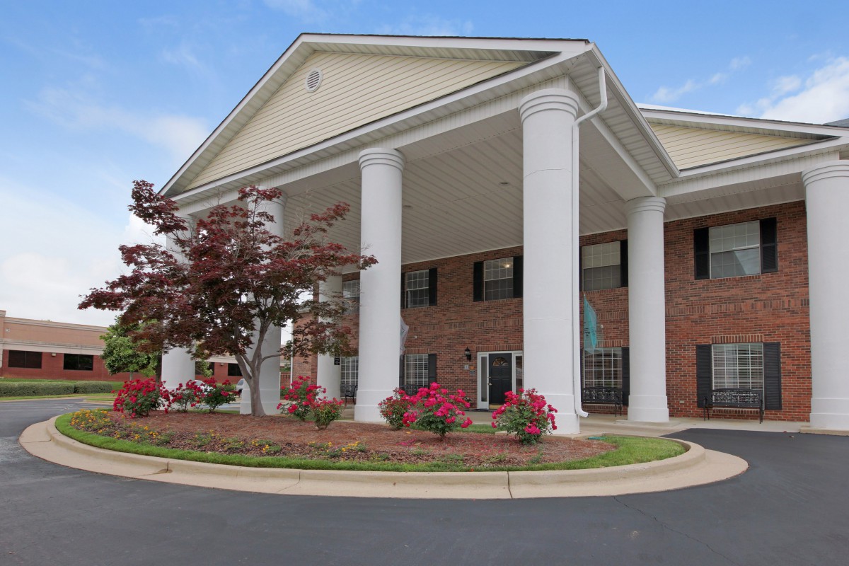 A building with large columns at its entrance, surrounded by landscaping and flowers under a blue sky.