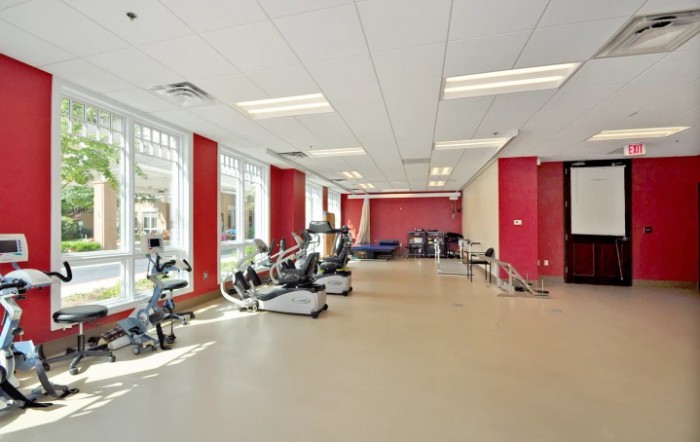 A spacious gym with red walls, equipped with treadmills and stationary bikes, well-lit by large windows.