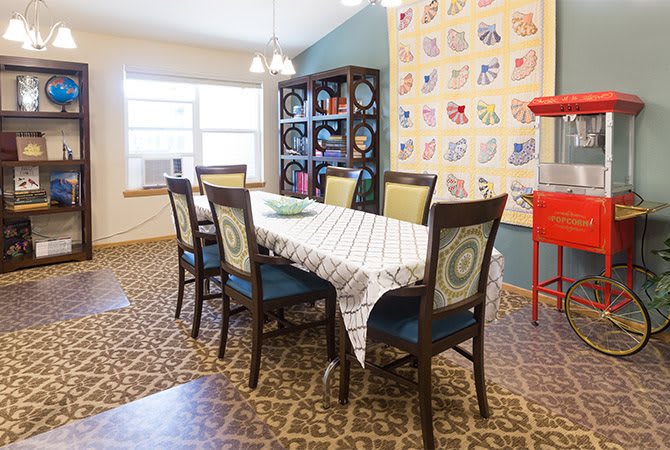 A cozy dining room with a table and six chairs, featuring a quilt on the wall and a bookshelf in the background.