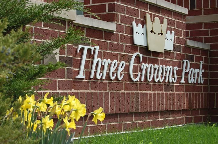 A brick wall featuring a sign that reads 'Three Crowns', with yellow daffodils in the foreground.