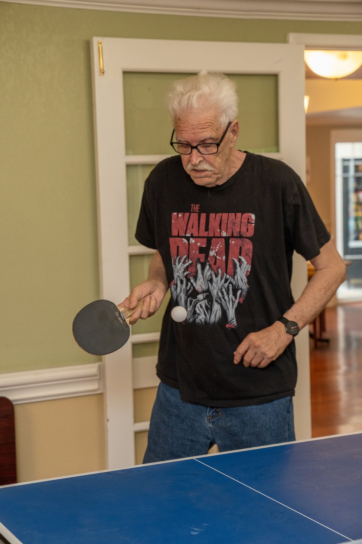 An elderly man playing table tennis indoors while wearing a casual t-shirt and glasses.