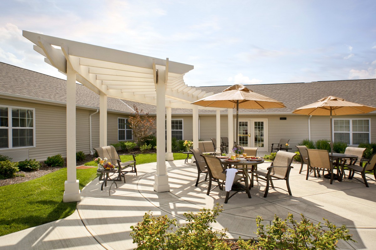 A sunny patio area with a pergola, featuring tables and chairs with umbrellas for shade.