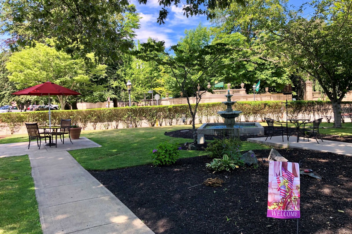 A serene garden with a decorative fountain, surrounded by lush greenery and a sunlit path.