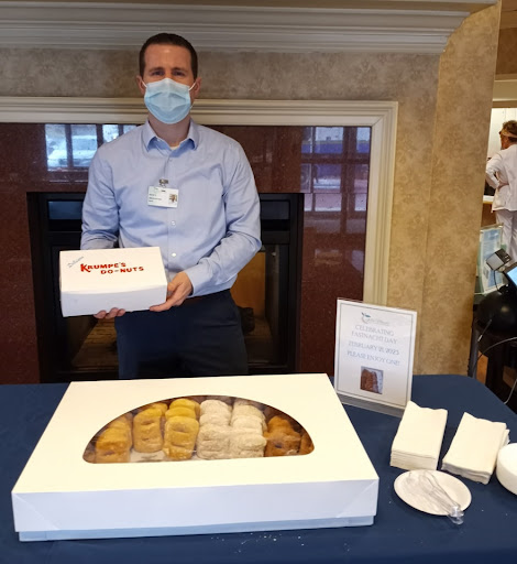 A man wearing a mask holds a box of cake while standing next to a table displaying an assortment of desserts.
