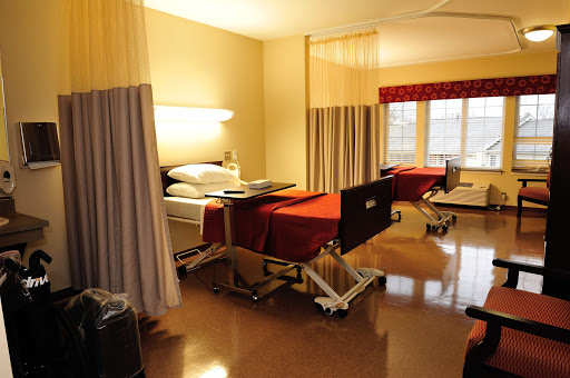 A view of a patient room in a hospital, featuring two beds with red covers and curtains for privacy.