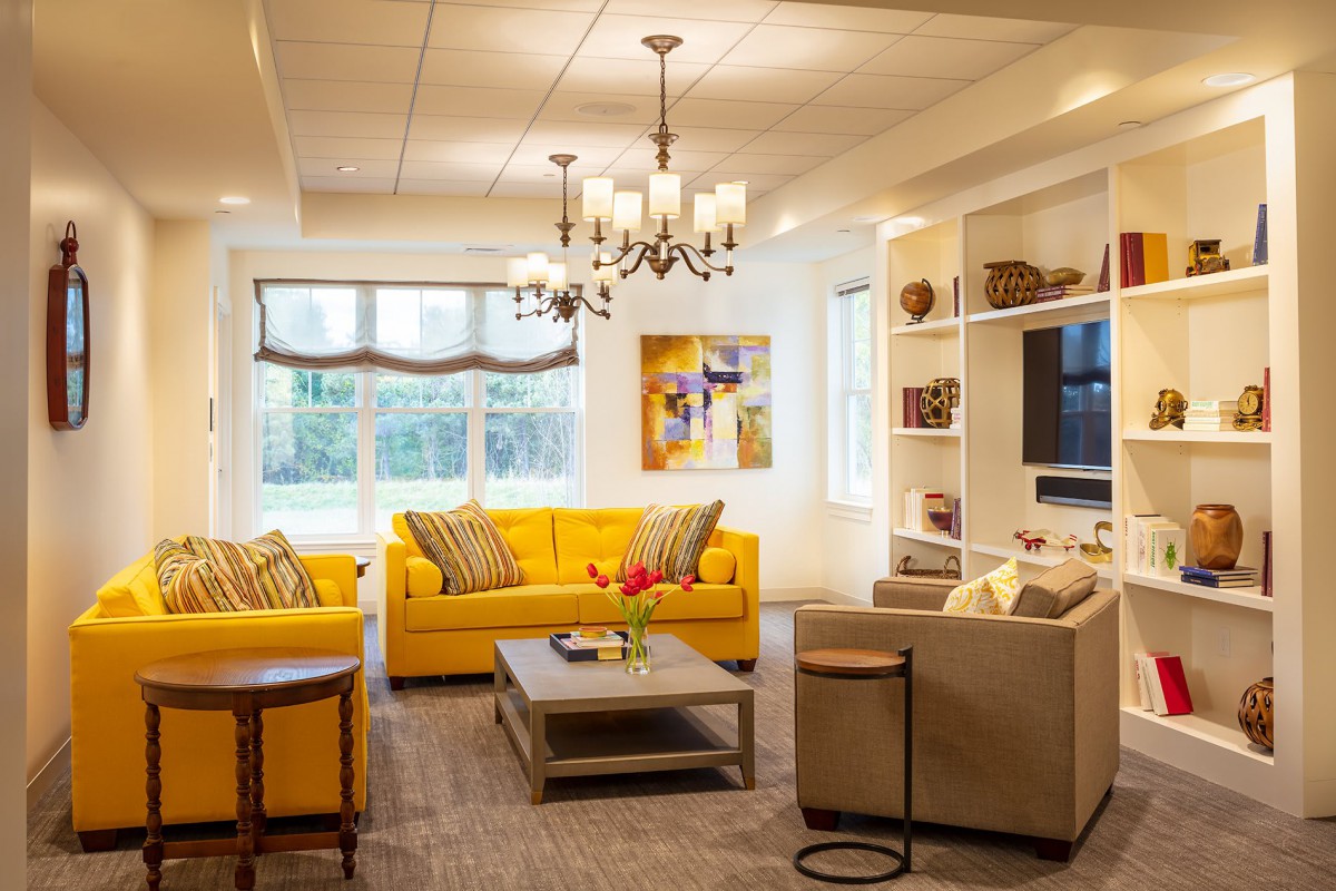 A bright and inviting living room featuring a yellow sofa, elegant chandelier, and neatly arranged bookshelf.