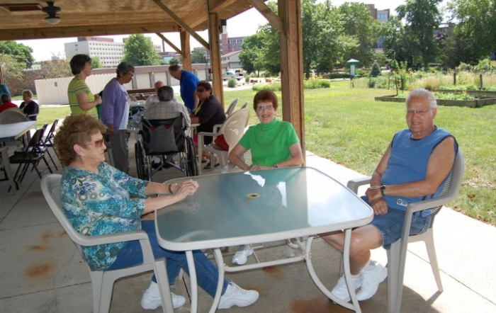 A group of people sitting around a glass table under a gazebo in a park.