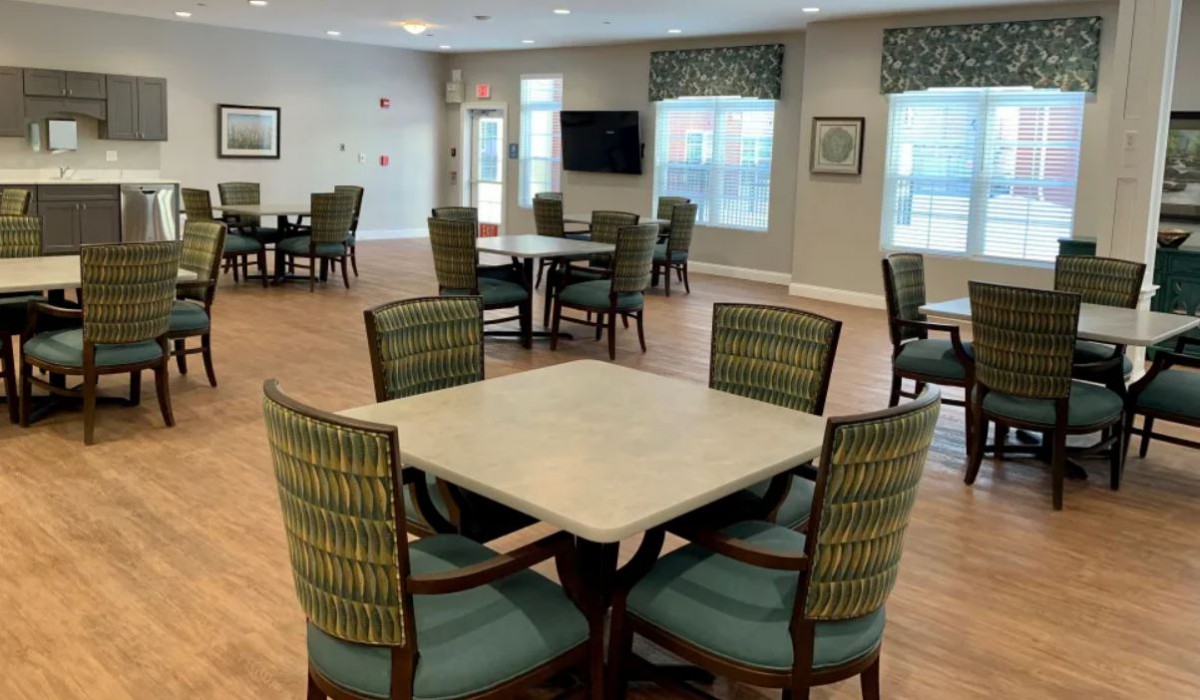 A spacious community dining area with several tables and chairs arranged neatly, featuring large windows and a wall-mounted television.