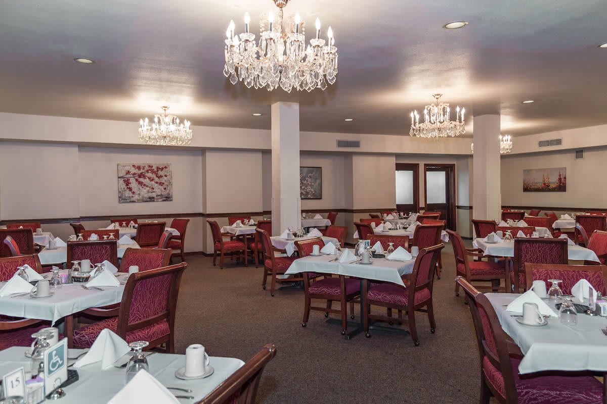 An elegant dining room with chandeliers, neatly arranged tables, and red upholstered chairs.