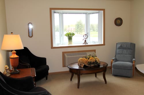 A cozy living room with a large bay window, comfortable chairs, and a wooden coffee table adorned with flowers.