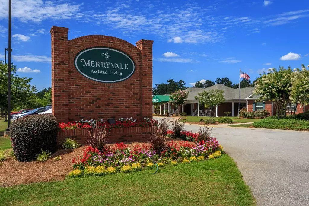 A beautifully landscaped entrance with a brick sign and vibrant flowers, leading to a nearby building under a partially cloudy sky.