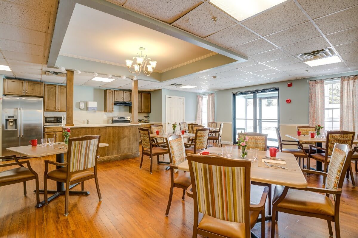 An empty dining room with wooden floors, several tables and chairs, and a kitchen area in the background.