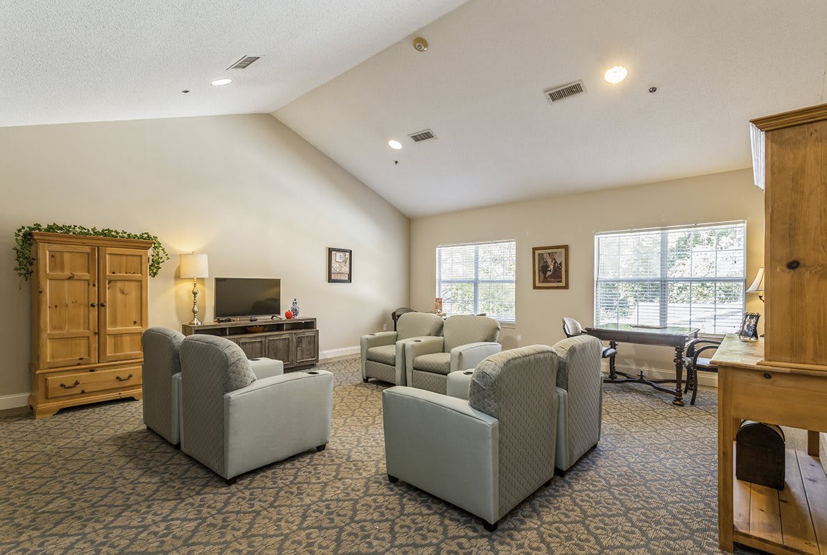 A cozy living room with armchairs arranged around a television, featuring large windows and carpeted flooring.