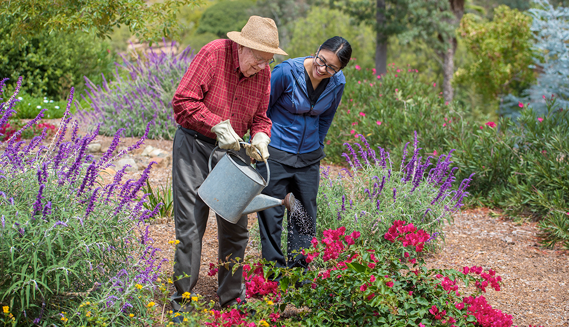 An elderly person waters flowers in a garden, assisted by another person.