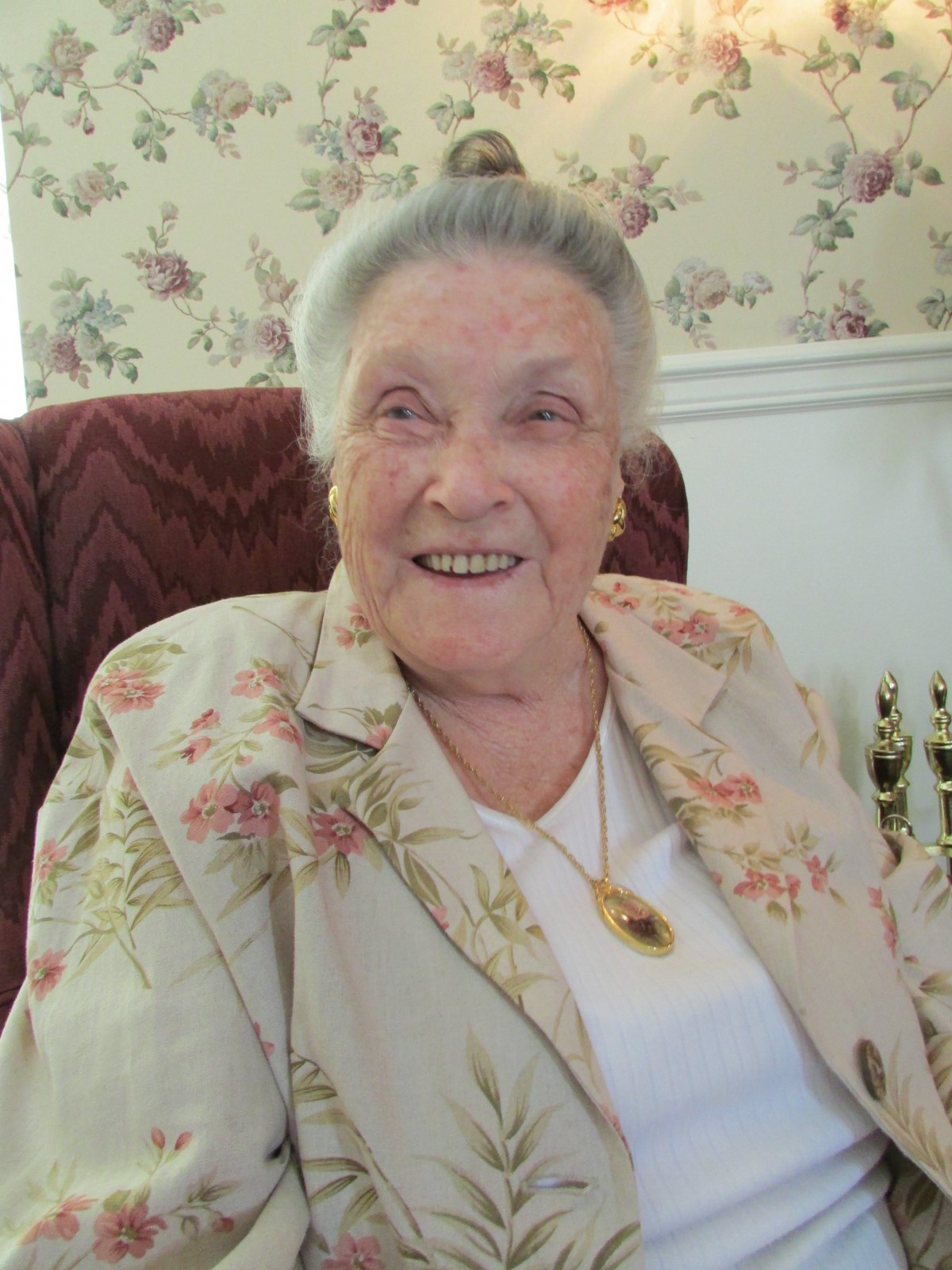 An elderly woman with a warm smile, wearing a floral jacket and necklace, seated in an armchair.