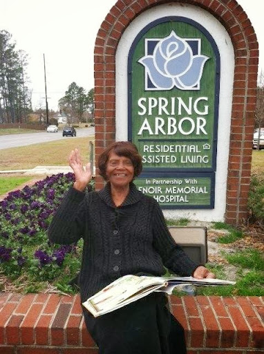 A woman happily waving while sitting in front of the Spring Arbor assisted living facility.