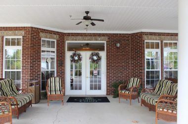 A welcoming brick porch with double doors adorned with wreaths, flanked by comfortable chairs and a ceiling fan.