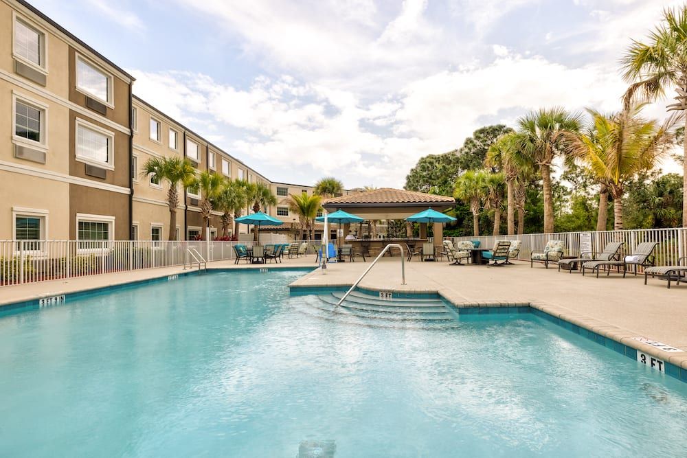 A sunny day at an outdoor hotel pool surrounded by palm trees with poolside chairs and umbrellas.