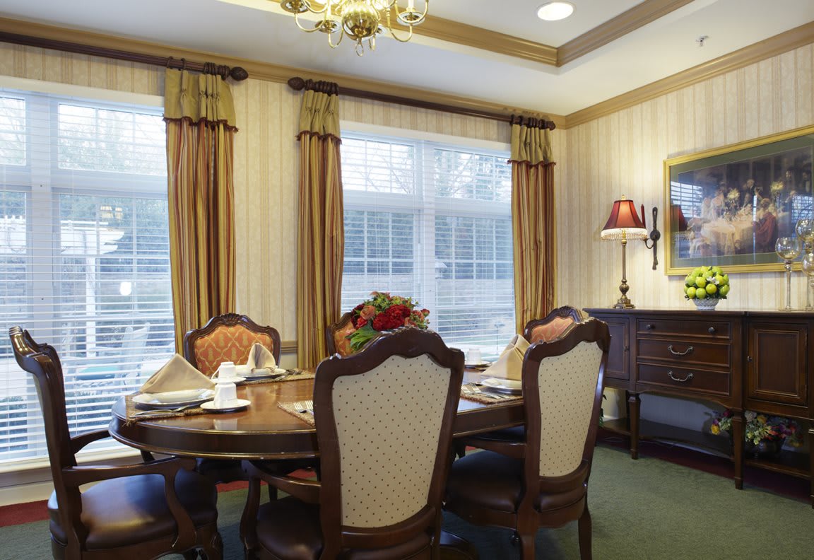 A classic dining room featuring a well-set table surrounded by upholstered chairs, with warm lighting from a chandelier and natural light streaming through the curtains.