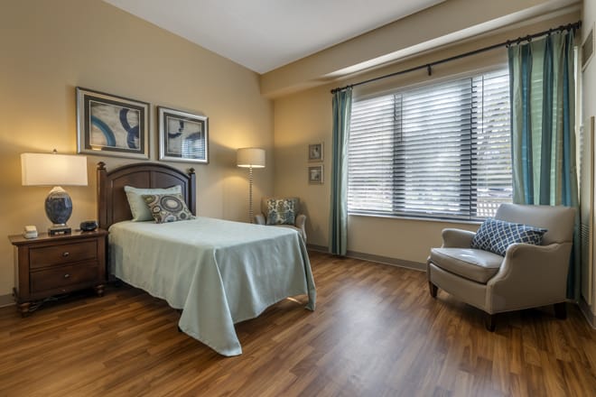 A cozy bedroom with a single bed, armchair, and large window with blinds.