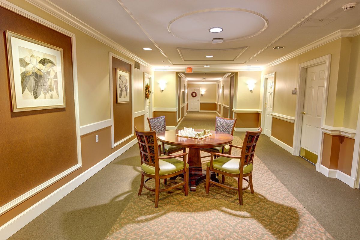 A well-lit hallway featuring a round table surrounded by chairs, with wall art and carpeted flooring.