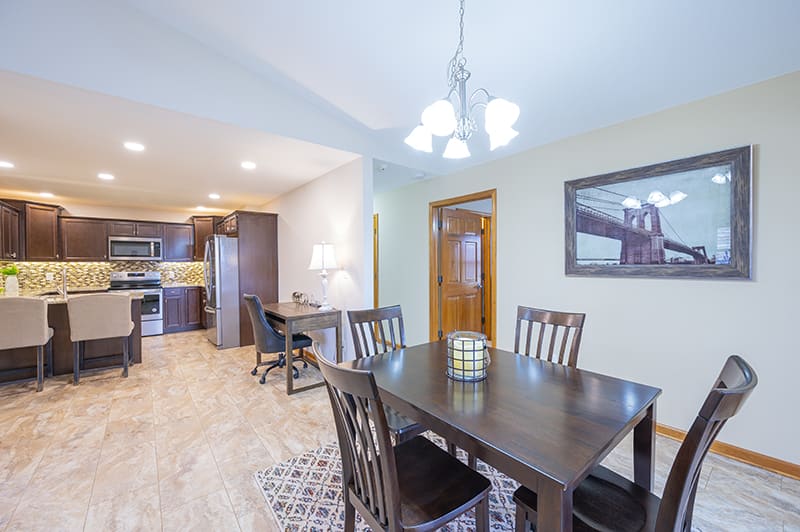 A modern dining room with a dark wooden table and chairs, adjacent to an open kitchen area, under a stylish light fixture.