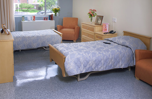 A bright and tidy hospital room featuring two patient beds, a cozy armchair, and decorative flowers by the window.