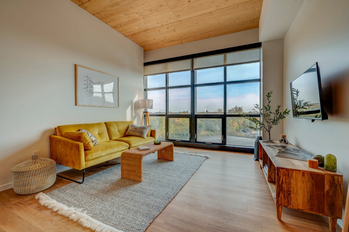 A modern living room with a yellow sofa, wooden ceiling, and large windows that bring in natural light.