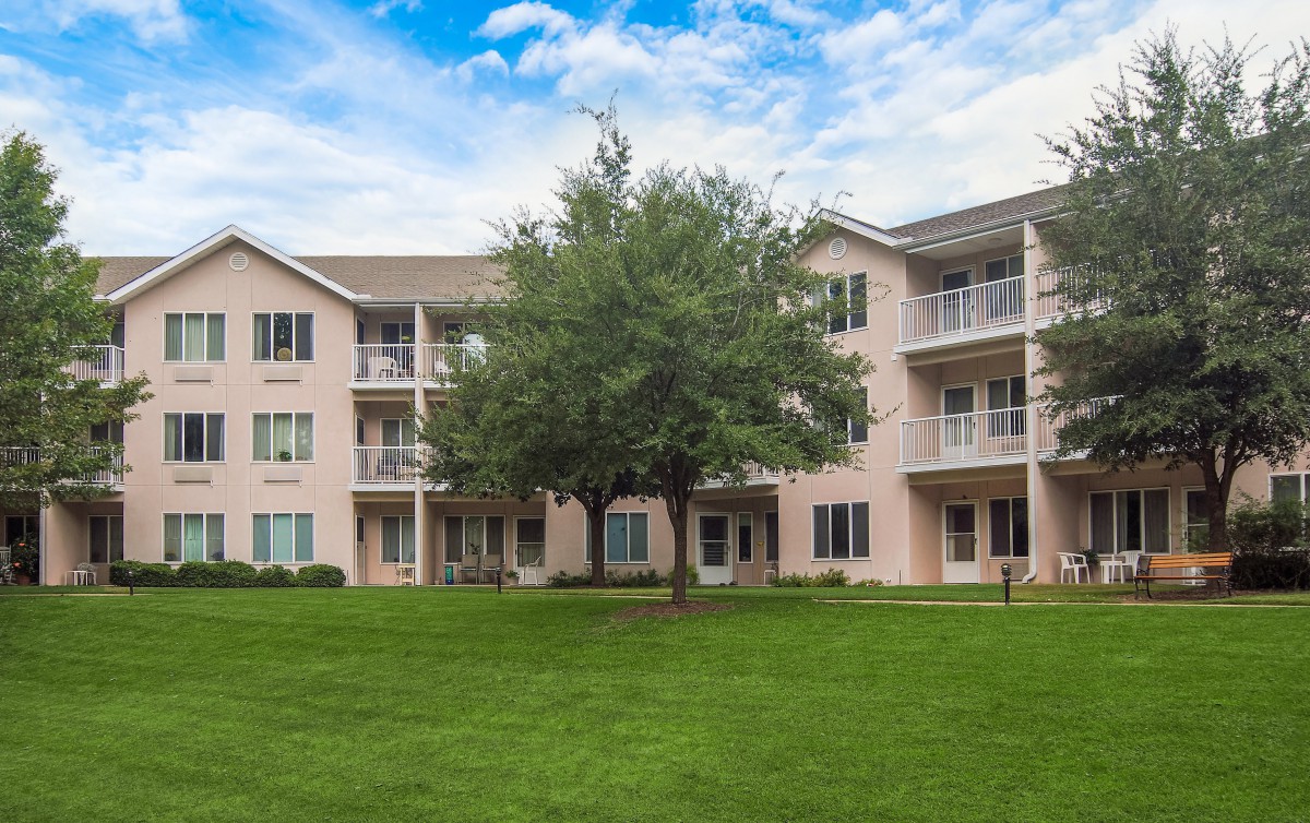 A multi-story residential apartment building with balconies, surrounded by a lush green lawn and trees.