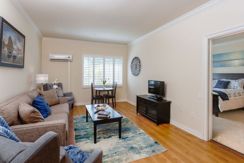 A cozy living room featuring a sofa, a television on a stand, a coffee table with decor items, and a small dining area near the window.