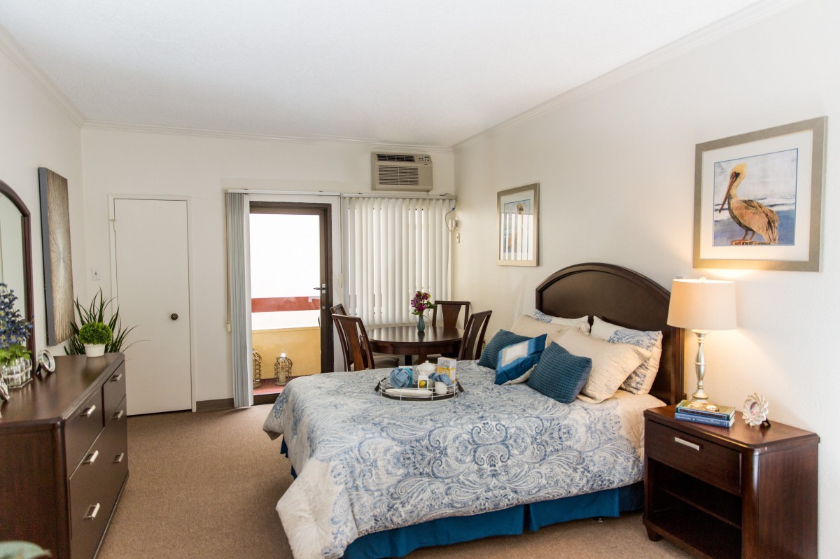 A cozy bedroom with a neatly made bed, decorated with blue accents, and a small dining area with a table and chairs near the window.