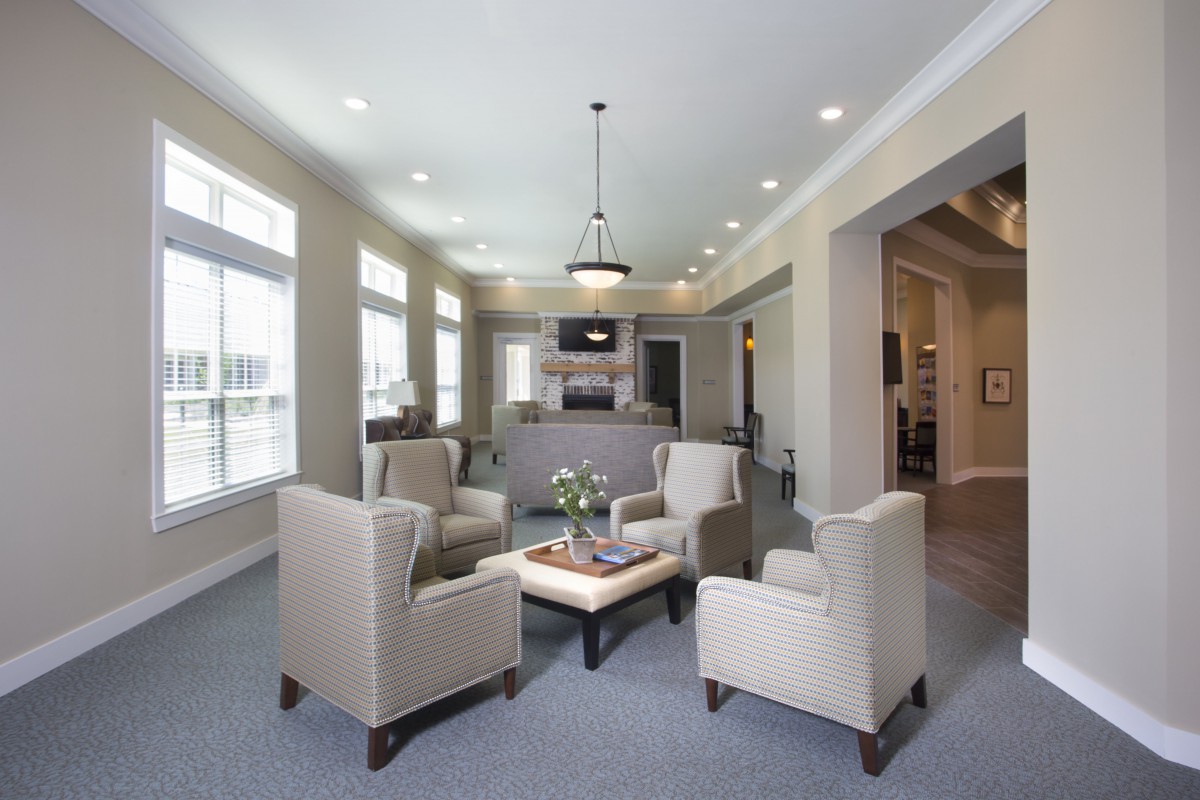 A modern living room with beige armchairs arranged around a table, featuring large windows and a view into the kitchen.