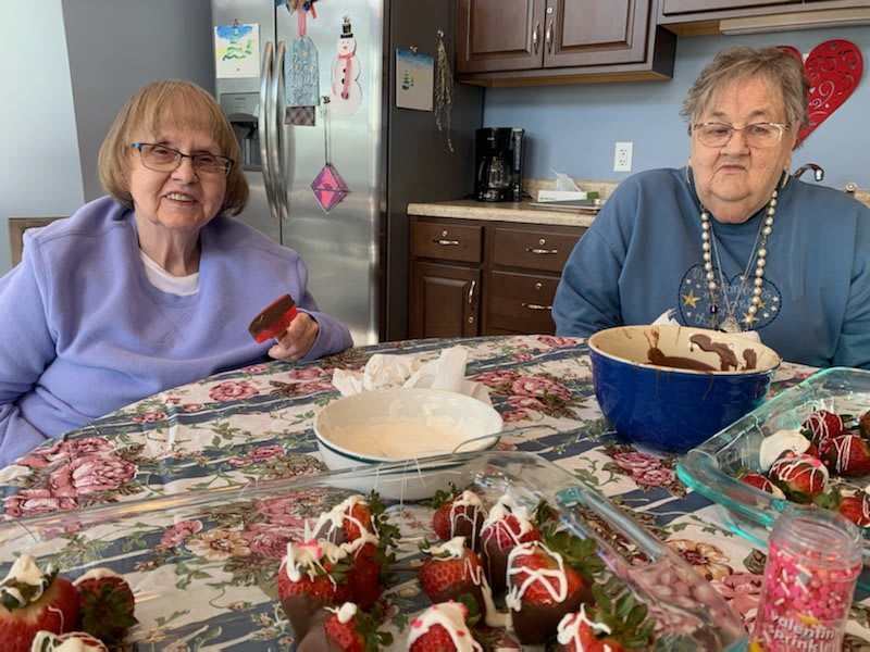 Two elderly women enjoy preparing desserts at a kitchen table.