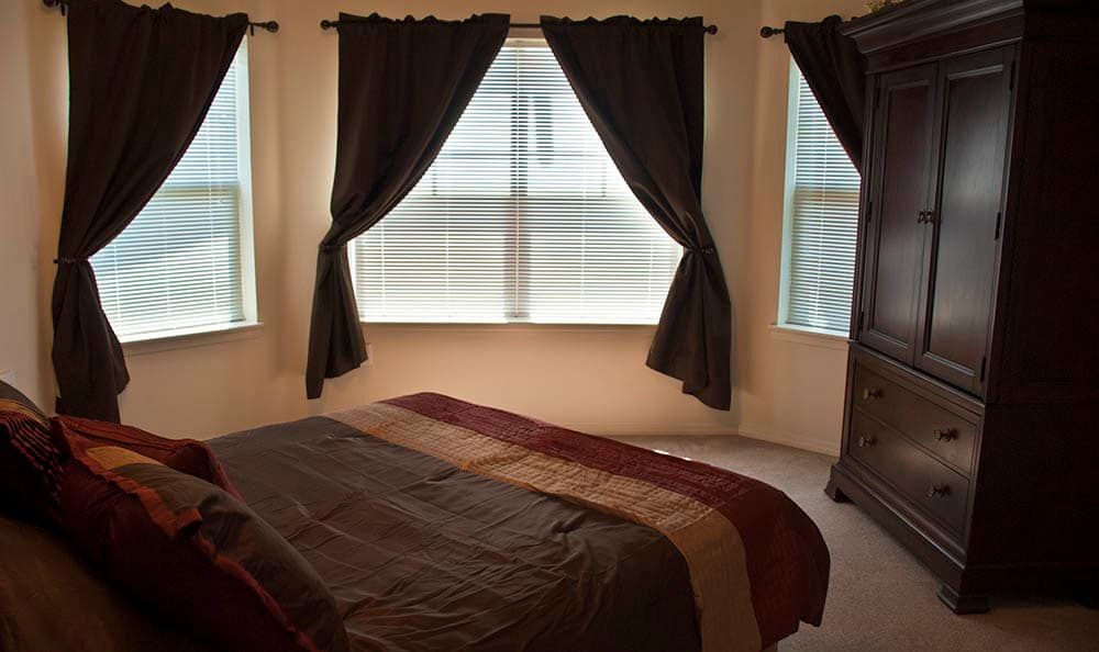 A cozy bedroom with a neatly made bed and large windows adorned with dark curtains.