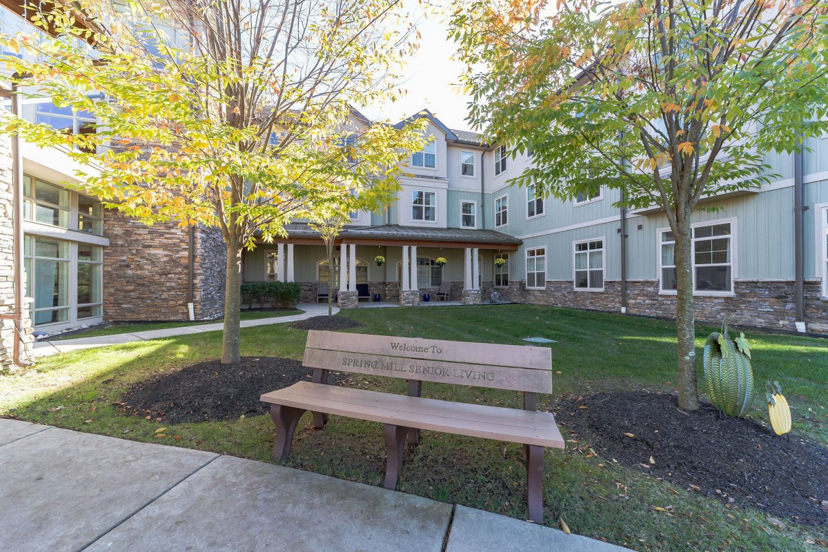 A peaceful outdoor area with a wooden bench in front of a residential building surrounded by trees and a well-maintained lawn.