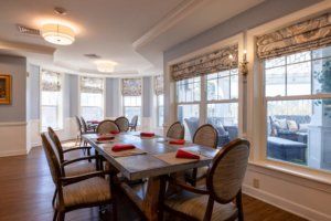 A well-lit dining room with a large table and neatly arranged chairs, accented by natural light streaming through the windows.