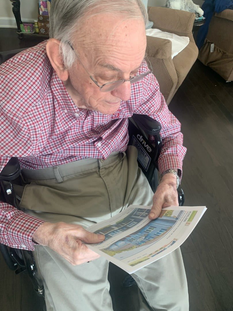 An elderly man seated in a wheelchair, reading a newspaper indoors.