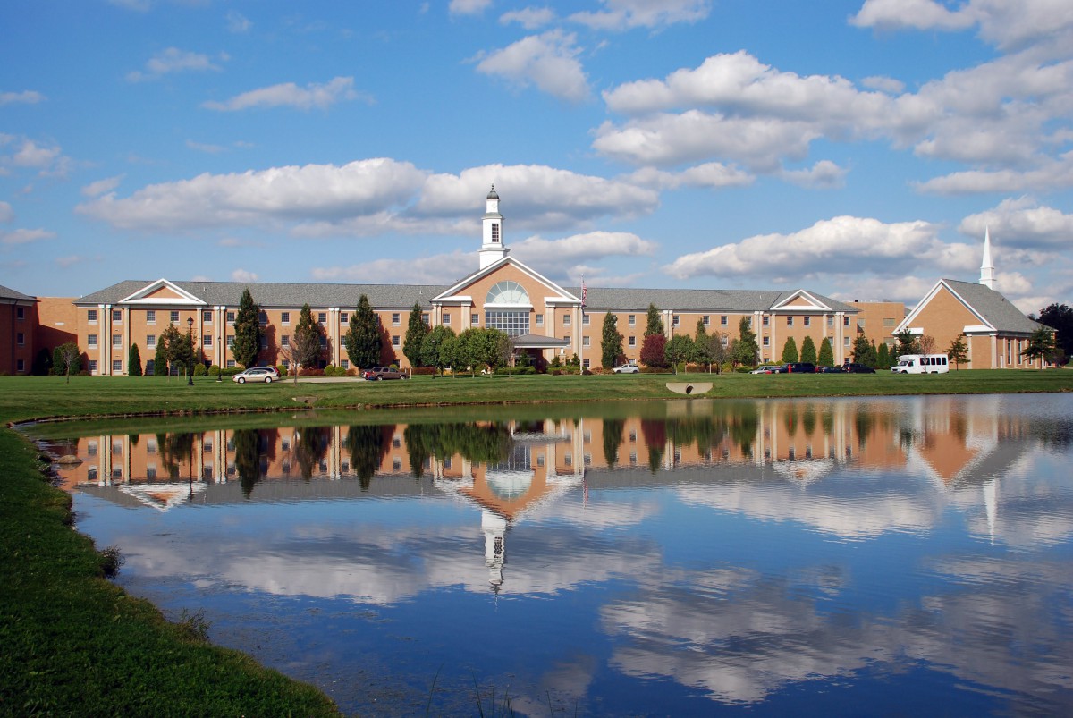 A large building with a spire reflecting in a calm lake under a partly cloudy sky.