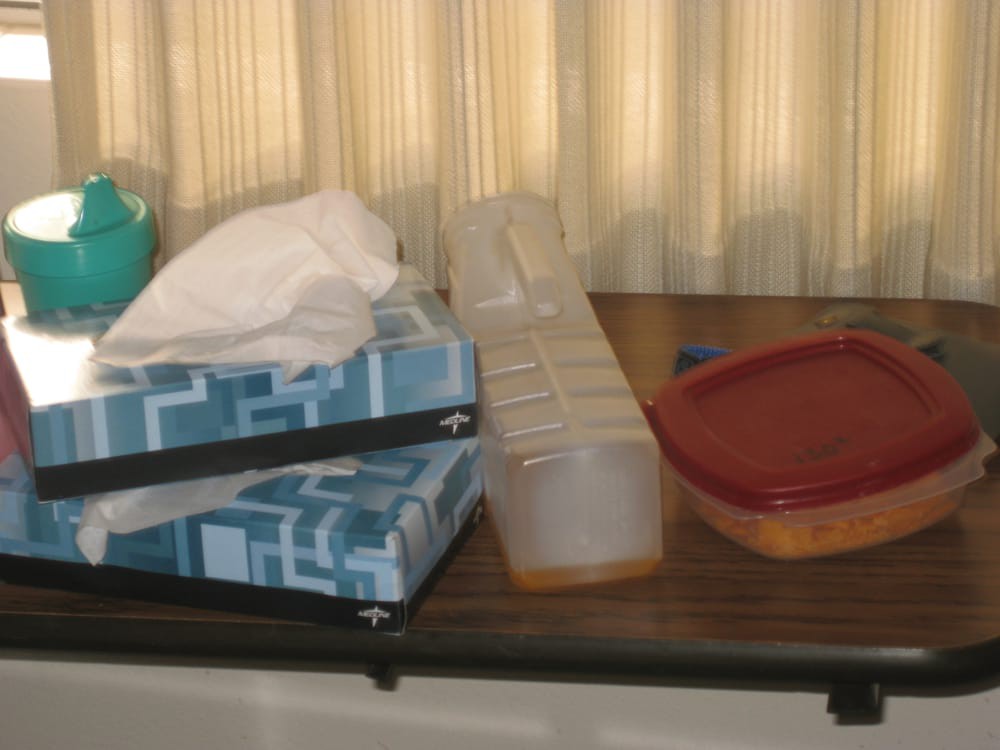 A table with a tissue box, plastic container, and lid in front of curtains.