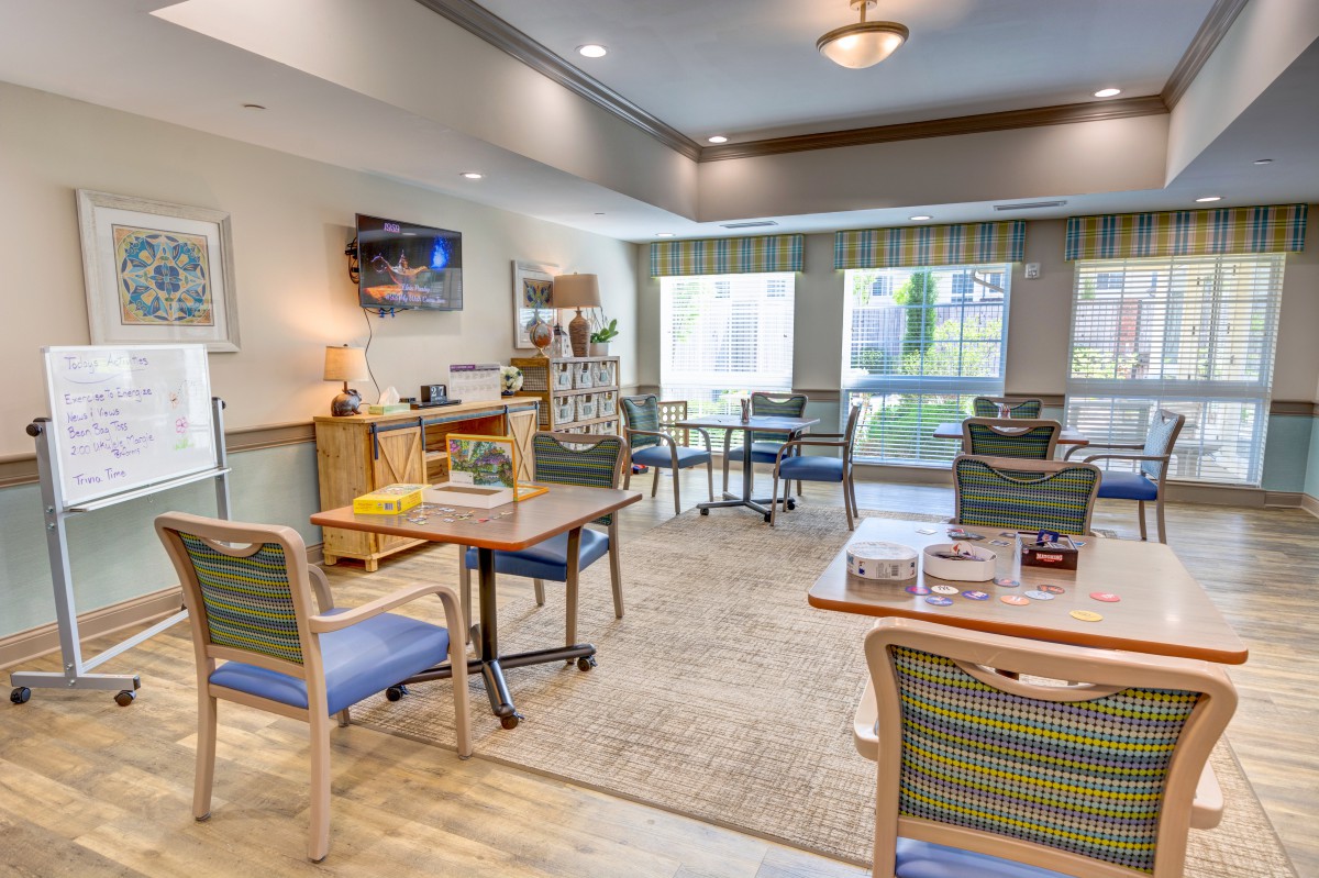 A cozy interior room with chairs, tables, and a television set, featuring large windows for natural light.