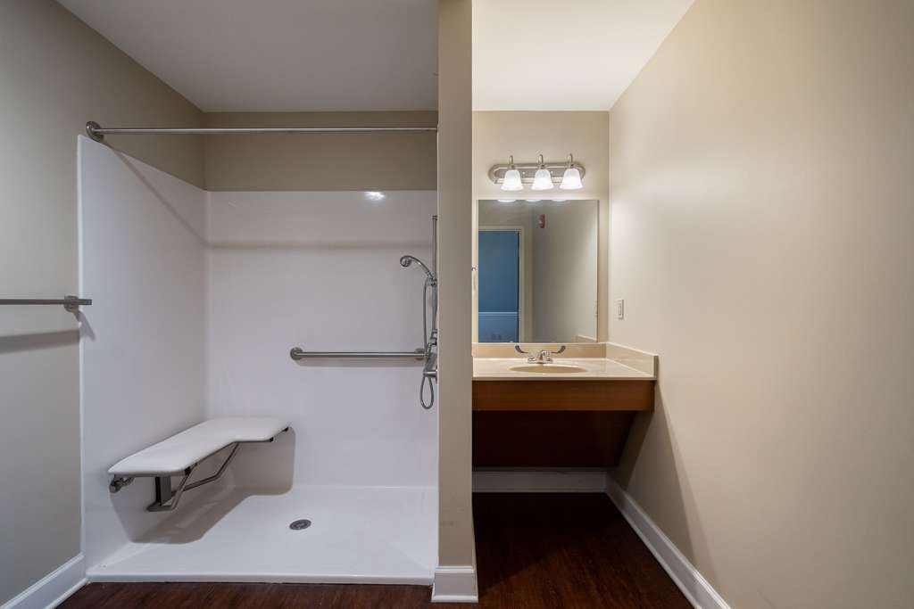 A bathroom featuring an accessible shower with a grab bar and foldable seat, alongside a vanity with a sink and mirror.